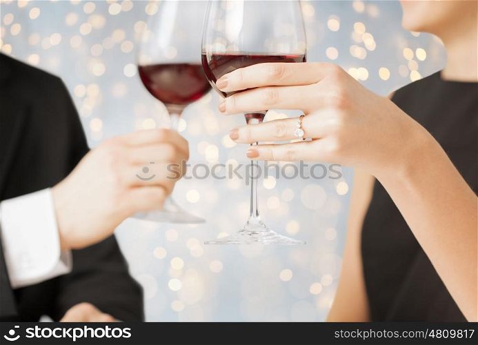 people, holidays, proposal, engagement and celebration concept - close up of engaged couple hands with ring and red wine glasses over lights background
