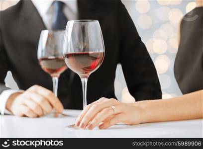 people, holidays, wedding, proposal and jewelry concept - hands of couple with diamond engagement ring and wine glasses in restaurant over lights background