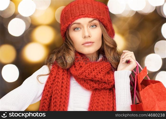 people, holidays, winter, christmas and sale concept - young woman in red woolen knitted hat and scarf holding shopping bags over lights background
