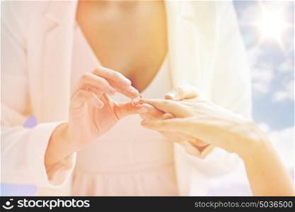 people, homosexuality, same-sex marriage and love concept - close up of happy lesbian couple hands putting on wedding ring over sky and sun background. close up of lesbian couple hands with wedding ring