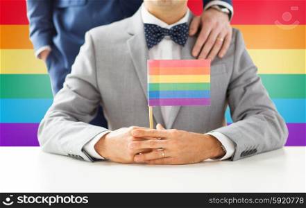 people, homosexuality, same-sex marriage and love concept - close up of happy male gay couple in suits and bow-ties with wedding ring holding rainbow flag over background