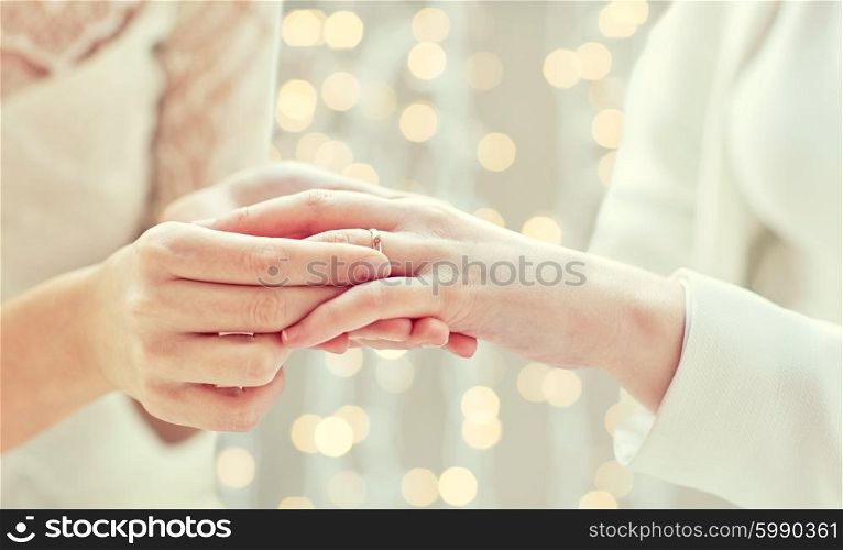 people, homosexuality, same-sex marriage and love concept - close up of happy lesbian couple hands putting on wedding ring over holidays lights background