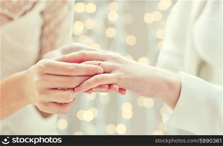 people, homosexuality, same-sex marriage and love concept - close up of happy lesbian couple hands putting on wedding ring over holidays lights background