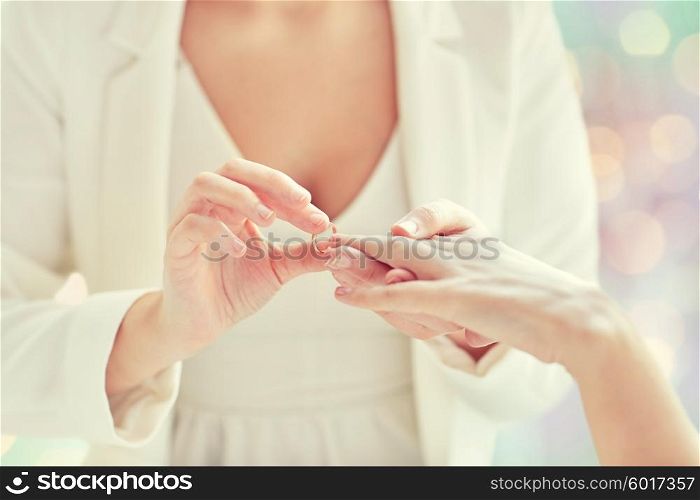 people, homosexuality, same-sex marriage and love concept - close up of happy lesbian couple hands putting on wedding ring over holiday lights background