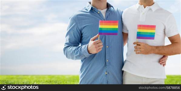 people, homosexuality, same-sex marriage, gay and love concept - close up of happy male gay couple hugging and holding rainbow flags over blue sky and grass background