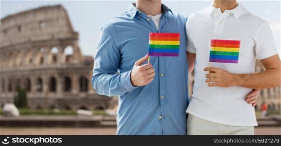 people, homosexuality, same-sex marriage, travel and love concept - close up of happy male gay couple holding rainbow flags and hugging from back over coliseum in rome background