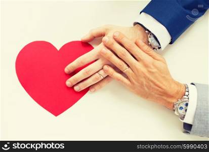 people, homosexuality, same-sex marriage, valentines day and love concept - close up of happy married male gay couple hands with red paper heart shape on table