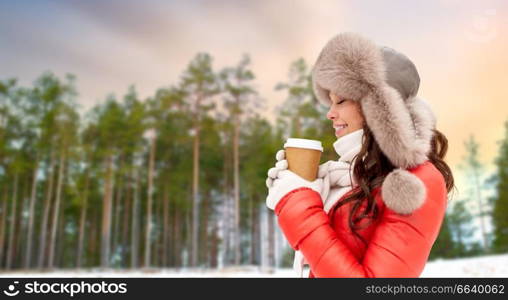 people, hot drinks and season concept - close up of happy woman in fur hat with coffee cup hat outdoors over winter forest background and snow. close up of woman with coffee cup in winter