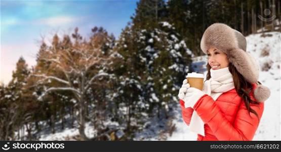 people, hot drinks and season concept - happy woman in fur hat with coffee cup hat outdoors over winter forest background. woman in fur hat with coffee over winter forest