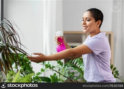 people, housekeeping and plants care concept - african american woman spraying houseplant with water sprayer at home. woman spraying houseplant with water at home