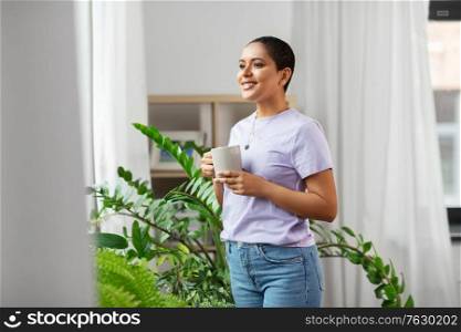 people, housekeeping and plants care concept - african american woman with houseplants drinking coffee at home. african american woman drinking coffee at home