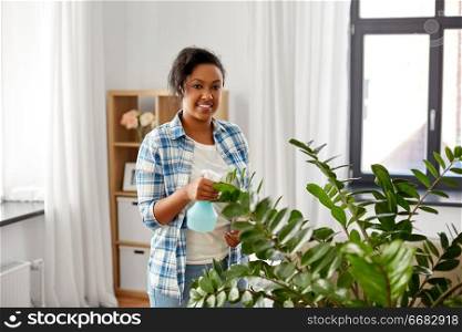 people, housework and care concept - happy african american woman or housewife spraying houseplant with water sprayer at home. happy woman spraying houseplant with water at home