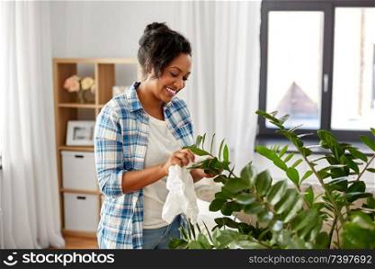people, housework and care concept - happy african american woman or housewife cleaning houseplant leaves by tissue at home. happy woman cleaning houseplant at home