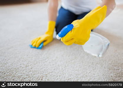 people, housework and housekeeping concept - close up of woman in rubber gloves with cloth and detergent spray cleaning carpet at home