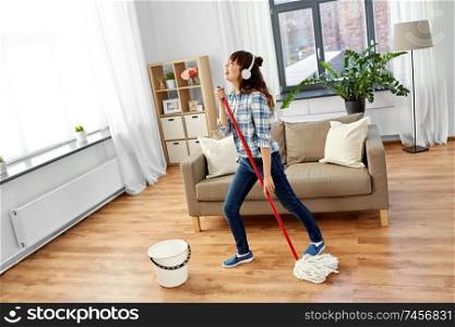 people, housework and housekeeping concept - happy asian woman or housewife in headphones with mop cleaning floor and singing at home. asian woman or housewife with mop singing at home