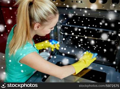 people, housework and housekeeping concept - happy woman with bottle of spray cleanser cleaning oven at home kitchen over snow effect