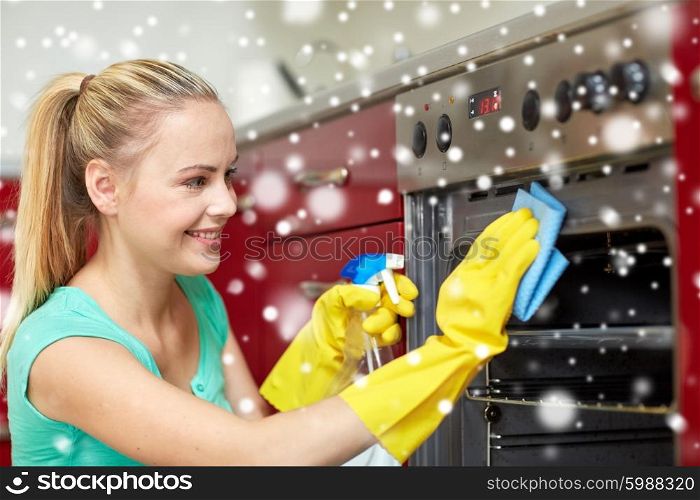 people, housework and housekeeping concept - happy woman with bottle of spray cleanser cleaning oven at home kitchen over snow effect