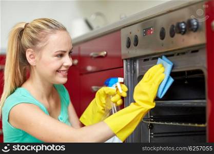 people, housework and housekeeping concept - happy woman with bottle of spray cleanser cleaning oven at home kitchen