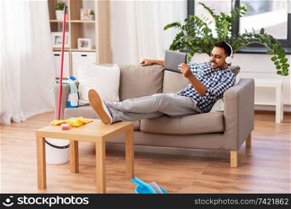 people, housework and housekeeping concept - indian man in headphones listening to music on tablet computer after home cleaning. indian man in headphones after cleaning home