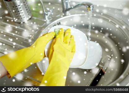 people, housework, washing-up and housekeeping concept - close up of woman hands in protective gloves washing dishes with sponge at home kitchen over snow effect