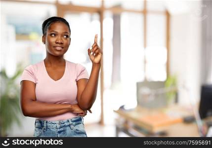 people, idea and attention concept - happy african american young woman pointing finger up over office background. happy african american woman pointing finger up