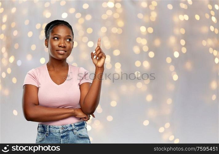 people, idea and holidays concept - happy african american young woman pointing finger up over festive lights background. happy african american woman pointing finger up