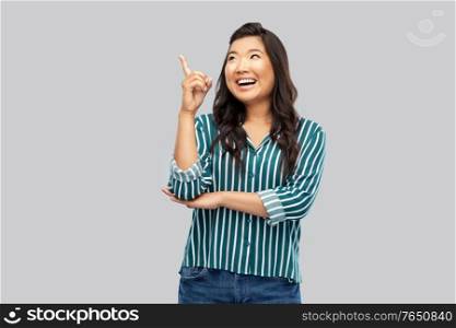 people, idea and inspiration concept - happy asian young woman pointing finger up over grey background. happy asian woman pointing finger up on grey