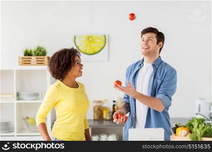 people, international and healthy eating concept - happy couple cooking food and juggling tomatoes at home kitchen. couple cooking food and juggling tomatoes at home