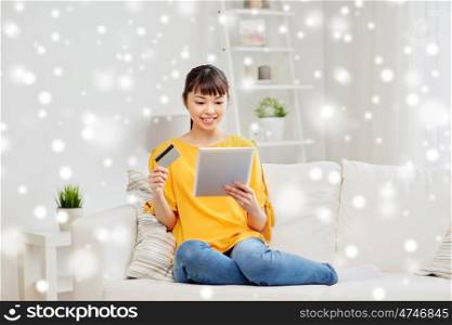 people, internet bank, online shopping, technology and e-money concept - happy asian young woman sitting on sofa with tablet pc computer and credit card at home over snow