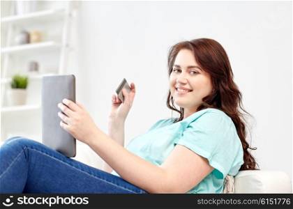 people, internet bank, online shopping, technology and e-money concept - happy young plus size woman sitting on sofa with tablet pc computer and credit card at home