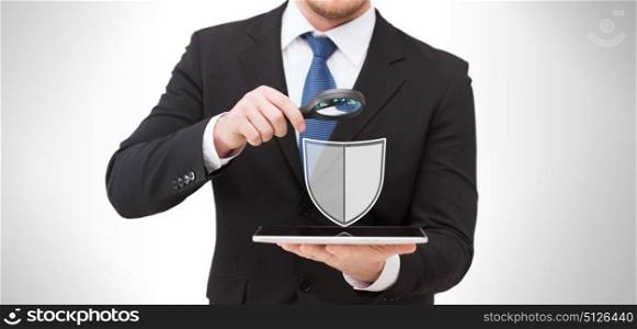 people, internet security and cyber protection concept - businessman holding magnifying glass over tablet pc computer with virtual antivirus program shield icon. businessman with tablet pc antivirus program icon