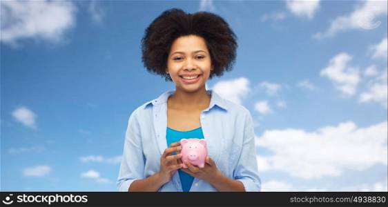 people, investment, saving and finances concept - happy afro american young woman with piggy bank over blue sky background. happy afro american young woman with piggy bank