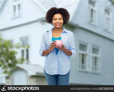 people, investment, saving and finances concept - happy afro american young woman with piggy bank over house background. happy afro american young woman with piggy bank