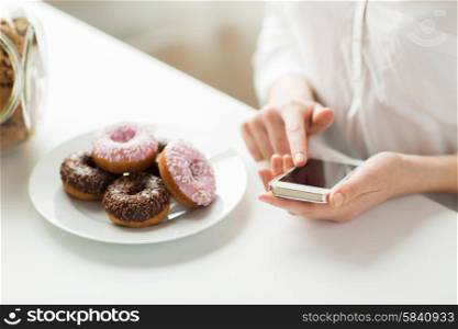 people, junk food, diet, technology and unhealthy eating concept - close up of hands with smart phone and donuts counting calories and sitting at table at home