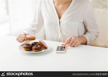 people, junk food, diet, technology and unhealthy eating concept - close up of hands with smart phone and donuts counting calories and sitting at table at home