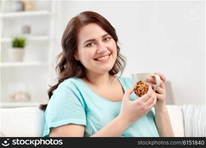 people, junk food, unhealthy eating and leisure concept - happy plus size young woman with cup of tea with cookie at home