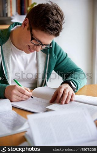 people, knowledge, education and school concept - happy student boy in glasses writing to notebook in library