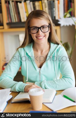 people, knowledge, education and school concept - happy student girl in eyeglasses with book and coffee cup in library