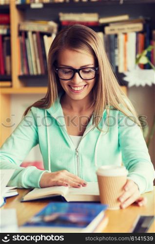 people, knowledge, education and school concept - happy student girl in eyeglasses reading book and drinking coffee in library