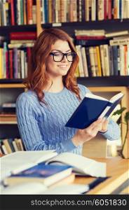 people, knowledge, education and school concept - happy student girl in eyeglasses reading book in library