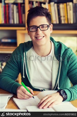 people, knowledge, education and school concept - happy student in eyeglasses with book writing to notebook in library