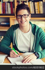 people, knowledge, education and school concept - happy student in eyeglasses with book writing to notebook in library