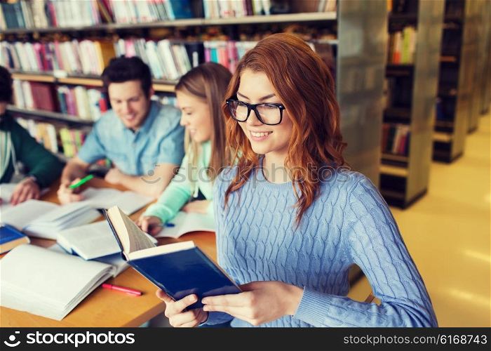 people, knowledge, education, literature and school concept - happy young woman in eyeglasses reading book and preparing to exams over group of students in library