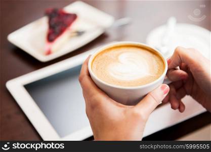 people, latte art and lifestyle concept - close up of hands with heart in coffee cup, tablet pc computer and cake on table