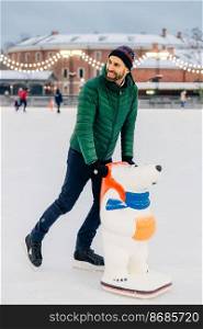 People, leisure, active lifestyle concept. Cheerful middle aged male learns to skate, uses skate aid, spends free time on skating rink, looks joyfully aside, enjoys spending weekends actively