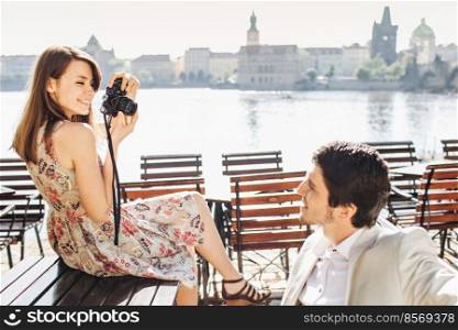 People, leisure and entertainment concept. Pleased beautiful female in summer dress males photos of her boyfriend, spend spare time together at outdoor cafeteria against wondeful landscapes.