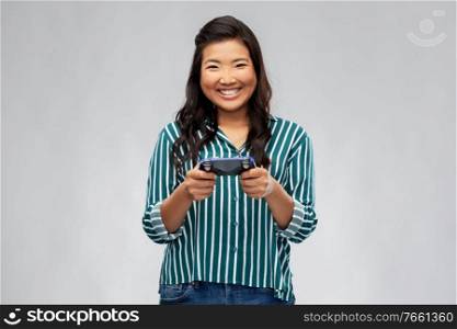 people, leisure and gaming concept - happy asian young woman playing video game with gamepad over grey background. happy asian woman playing video game with gamepad