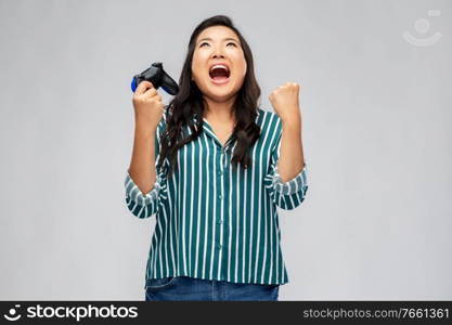 people, leisure and gaming concept - happy asian young woman playing video game with gamepad celebrating success over grey background. happy asian woman playing video game with gamepad