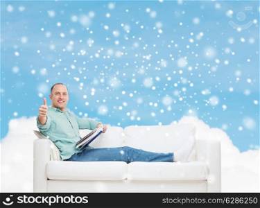 people, leisure and happiness concept - smiling man with book lying on sofa and showing thumbs up over blue sky with cloud and snow background