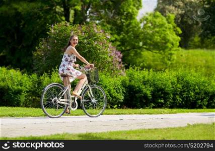 people, leisure and lifestyle - happy young hipster woman wearing summer dress riding fixie bicycle with wild flowers in basket at park. happy woman riding fixie bicycle in summer park